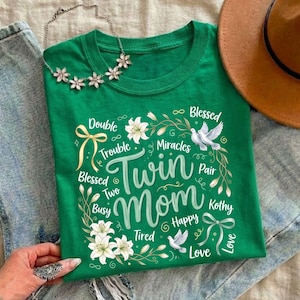 May include: A green t-shirt with the words "Twin Mom" in a cursive font, surrounded by floral and dove illustrations. The shirt also features the words "Double Trouble", "Blessed", "Miracles", "Happy", and "Love".