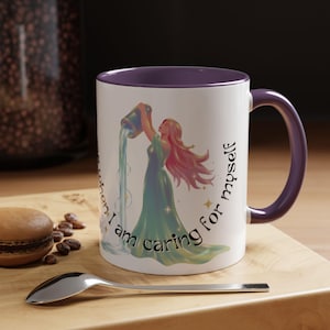 May include: White ceramic mug with a purple interior and handle. The mug features an illustration of a woman pouring water from a jug, with the text "When I am caring for myself". A spoon and a macaron are in the foreground.