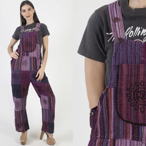 Op de afbeelding: Paarse patchwork tuinbroek met geborduurde bloemmotieven. De tuinbroek heeft een borstzak en verstelbare bandjes. De stof is een mix van gestreepte en effen paarse stoffen. Het model draagt er een grijs t-shirt onder.