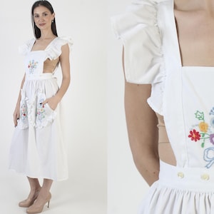 May include: A white apron dress with embroidered floral details. The dress features ruffled sleeves and pockets, with a square neckline and a midi-length skirt. The embroidery includes red, blue, yellow, and purple flowers.