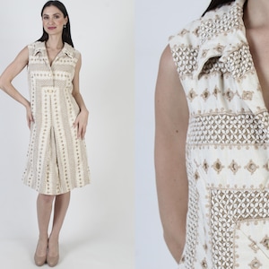 May include: A sleeveless, knee-length dress in cream and brown. The dress features a collared neckline, button-down front, and a pleated skirt. The fabric has a repeating geometric pattern. The model is wearing beige heels.