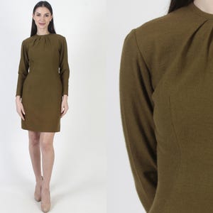 Puede incluir: Un vestido largo de manga larga, de color verde oliva, con cuello alto y un detalle plisado en el cuello.