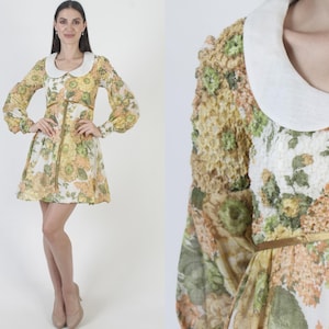 Pode incluir: Um vestido curto com base branca e padrão floral amarelo e verde, gola redonda branca lisa e uma fita dourada fina atada na cintura. O vestido tem mangas compridas com punhos e um tecido com aspeto texturado e em camadas.
