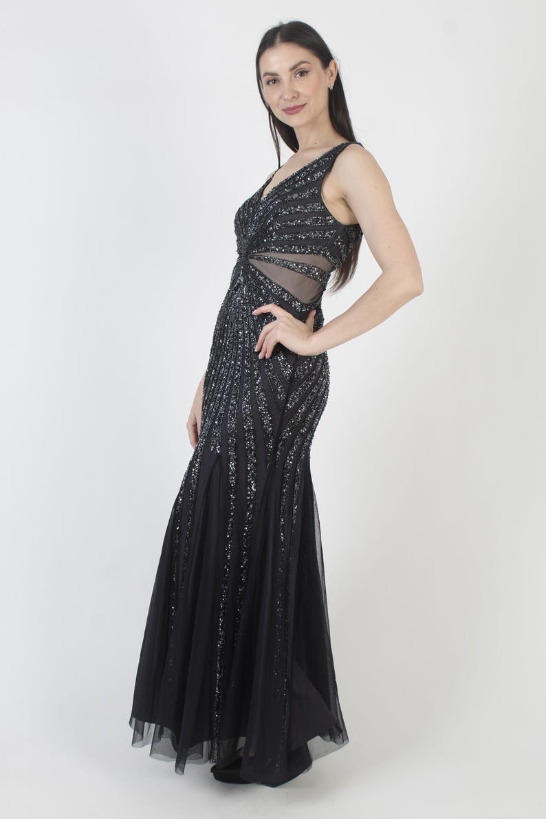 May include: A sleeveless black evening gown with a V-neckline and a flowing skirt. The dress is embellished with vertical rows of silver sequins. The bodice features a sheer panel at the waist. The gown is floor-length and suitable for formal events.