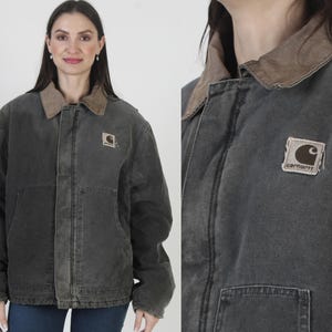 May include: A dark gray jacket with a brown corduroy collar. The jacket has a square logo patch on the chest and two large pockets. The jacket is worn over a dark shirt and blue jeans.