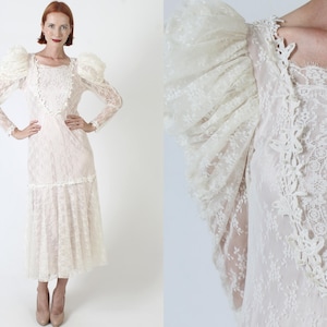 May include: A white lace wedding dress with a fitted bodice and a full skirt. The dress has long sleeves with puffed shoulders and a lace trim around the neckline.