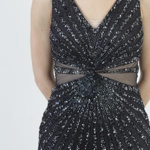 May include: A sleeveless black evening gown with a V-neckline and a fitted silhouette. The dress is heavily embellished with silver sequins and beads arranged in vertical and radial patterns, creating a shimmering effect. The waist features a sheer panel.