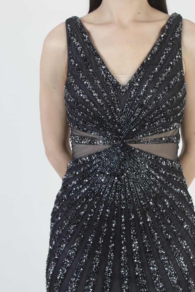 May include: A sleeveless black evening gown with a V-neckline and a fitted silhouette. The dress is heavily embellished with silver sequins and beads arranged in vertical and radial patterns, creating a shimmering effect. The waist features a sheer panel.