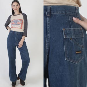 May include: A woman wearing a beige and gray baseball-style shirt with a Budweiser logo and dark blue high-waisted jeans. The jeans have a pocket with a small label that reads "Rodeo Bros."