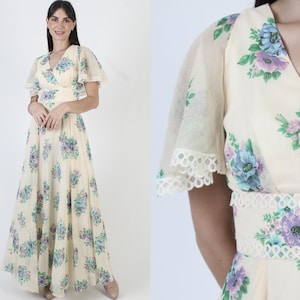 May include: A cream-colored, vintage-style maxi dress with a floral pattern in shades of blue, purple, and green. The dress features a V-neckline, short, sheer sleeves with lace trim, and a fitted waist. The skirt flows to the floor.