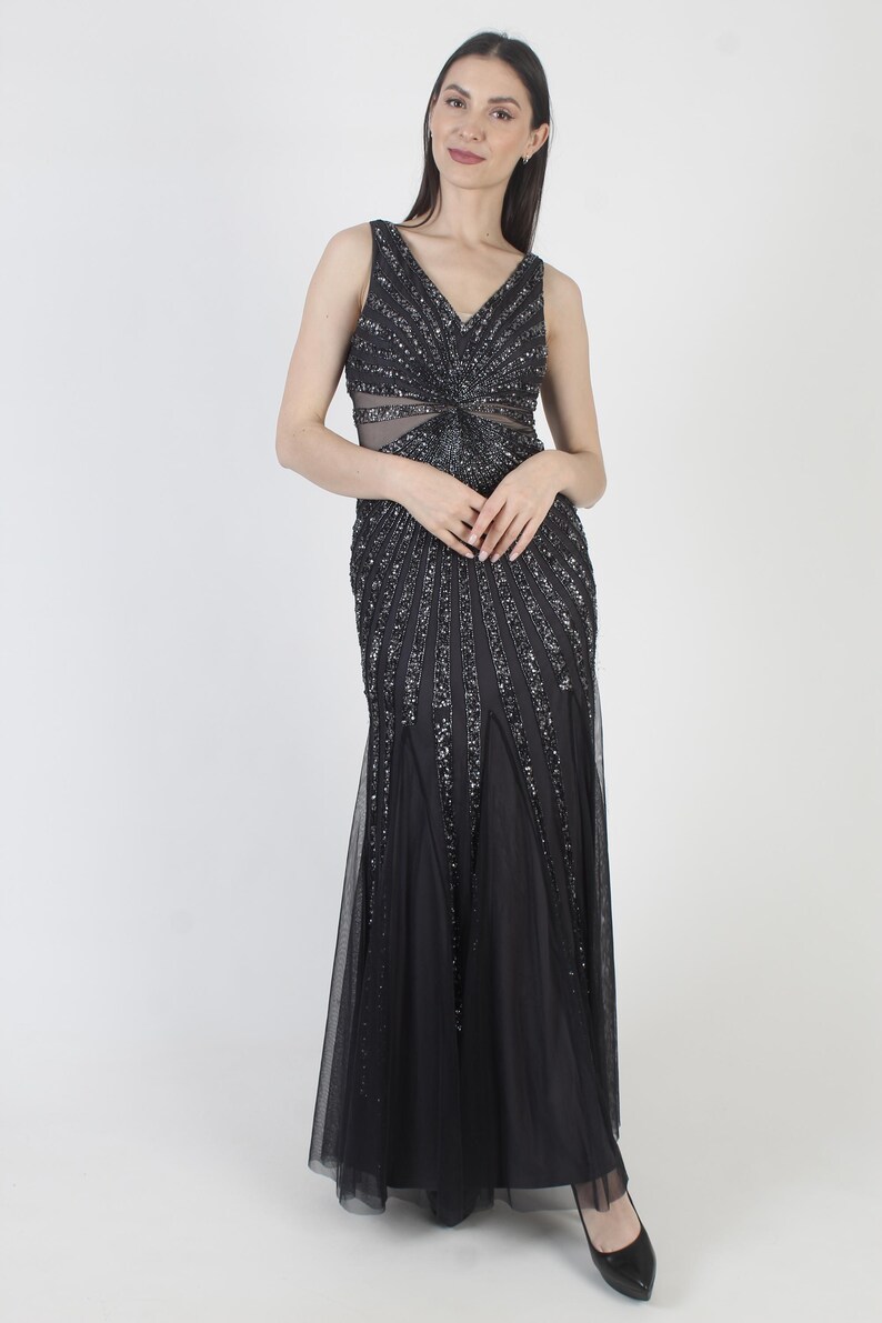 May include: A sleeveless black evening gown with a V-neckline and a flowing skirt. The bodice and upper skirt are embellished with sparkling sequins, creating a radiant effect. The dress features a sheer panel at the waist.