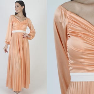 Peut inclure: Une longue robe en satin pêche avec un corsage plissé et une jupe plissée. La robe a des manches longues et une ceinture blanche.