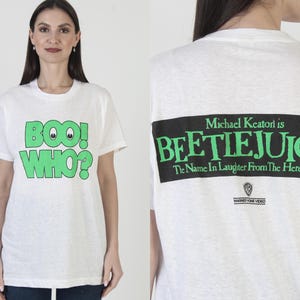 May include: White t-shirt with green text that says "BOO! WHO?" on the front. The back of the shirt has a black rectangle with the words "Michael Keaton is BEETLEJUICE" in green, and "The Name In Laughter From The Hereafter" in white.