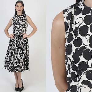 May include: Sleeveless black and white floral print dress. The dress features a high neckline, fitted bodice, and a flared skirt that falls to the mid-calf. The black floral pattern is set against a white background. The model is wearing black heels.