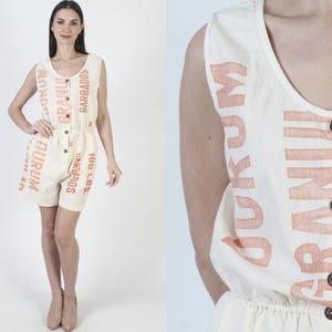 May include: A white sleeveless jumpsuit with a drawstring waist and a button closure. The jumpsuit is printed with the words "GRANUM DURUM" and "BARBADOS" in orange.