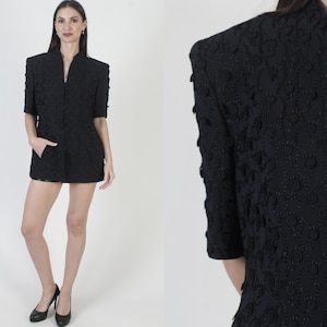May include: Black blazer with a textured floral pattern, short sleeves, and a stand-up collar. The blazer has a zipper closure and two front pockets. The model is wearing black heels. The blazer is a vintage style.