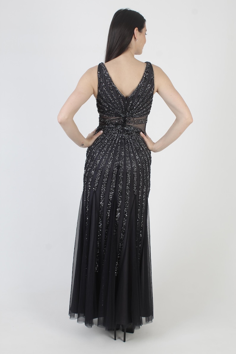 May include: A black formal gown with a V-neckline and a flowing skirt. The bodice and upper skirt are embellished with vertical rows of sequins. The dress features a sheer panel at the waist and a tulle overlay.