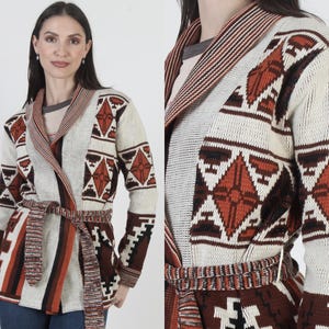 Könnte beinhalten: Cremefarbene Strickjacke mit geometrischen Mustern in Braun-, Schwarz- und Orangetönen. Der Pullover hat einen Schalkragen und einen passenden Gürtel. Das Design umfasst Diamanten- und Rechteckformen und schafft eine südwestliche oder Stammesästhetik.