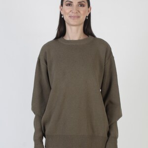Puede incluir: Un su&eacute;ter verde oliva con cuello redondo y mangas largas. El su&eacute;ter tiene un dobladillo y pu&ntilde;os acanalados. La modelo lleva vaqueros azules. El fondo es blanco liso.