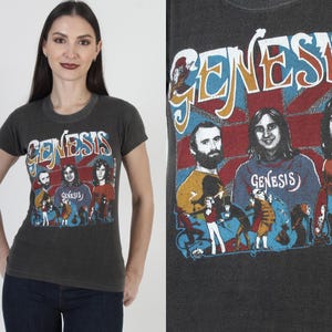 May include: A black t-shirt with a vintage-style graphic of the band Genesis. The graphic features the band's name in a colorful, stylized font and includes images of the band members.