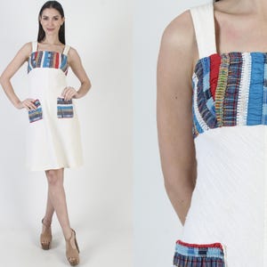 May include: A white sleeveless dress with a colorful geometric pattern on the bodice and pockets. The dress has a square neckline and a straight skirt.
