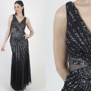 May include: A black formal gown with a V-neckline and intricate sequin and bead embellishments. The dress features a fitted bodice with a sheer midriff and a flowing, floor-length skirt. The design includes a detailed, radiating pattern.