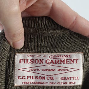 Puede incluir: Primer plano de una prenda de punto marr&oacute;n con una etiqueta que dice "FILSON GARMENT". La etiqueta tambi&eacute;n indica "100% Virgin Wool" y "C.C. FILSON CO. - SEATTLE". La prenda es de limpieza en seco profesional.