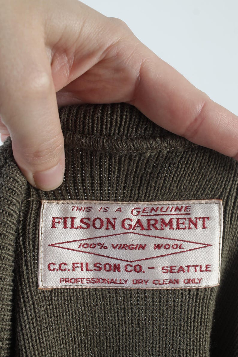 Puede incluir: Primer plano de una prenda de punto marr&oacute;n con una etiqueta que dice "FILSON GARMENT". La etiqueta tambi&eacute;n indica "100% Virgin Wool" y "C.C. FILSON CO. - SEATTLE". La prenda es de limpieza en seco profesional.