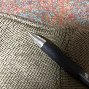 Puede incluir: Primer plano de un bol&iacute;grafo negro con punta plateada sobre tejido de punto verde oliva. El tejido tiene un patr&oacute;n acanalado texturizado. El fondo presenta un patr&oacute;n abstracto colorido.