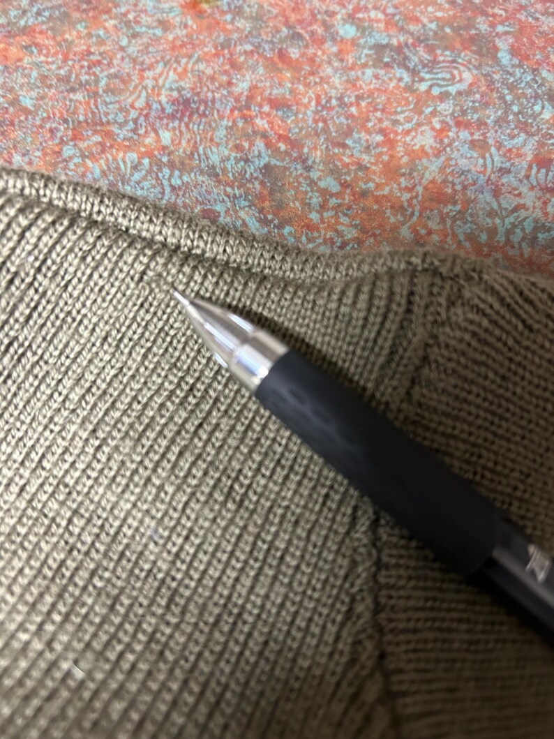 Puede incluir: Primer plano de un bol&iacute;grafo negro con punta plateada sobre tejido de punto verde oliva. El tejido tiene un patr&oacute;n acanalado texturizado. El fondo presenta un patr&oacute;n abstracto colorido.