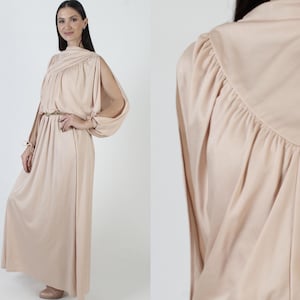 Peut inclure: Une longue robe maxi fluide de couleur beige clair. La robe a un col drapé et des manches longues avec des découpes aux épaules. La robe est cintrée à la taille par une fine ceinture dorée.