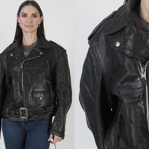 May include: A black leather jacket with a notched collar, zipper closure, and a belt at the waist. The jacket has multiple pockets and silver hardware. The model is wearing blue jeans.