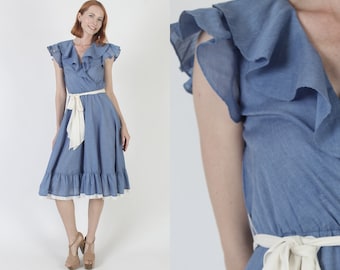 Puede incluir: Un vestido de mezclilla azul claro con ribete de volantes blancos y un lazo blanco atado a la cintura.