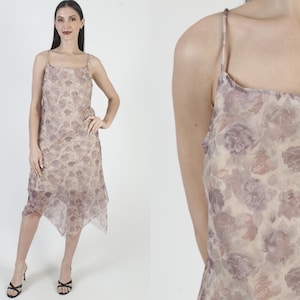 May include: A floral print slip dress in shades of beige, mauve, and purple. The dress features thin spaghetti straps and a midi length with an asymmetrical hemline. The fabric appears sheer and lightweight, suitable for warm weather.