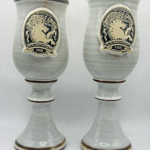 May include: Two tall, light gray ceramic goblets with a speckled finish and brown rim accents. Each features a raised circular emblem depicting a unicorn and the text Minnesota Renaissance Festival 15th Anniversary 1985 on the front surface.