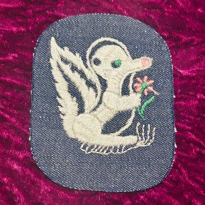 May include: A rounded rectangular denim patch featuring an embroidered white creature with a long snout, a green eye, and a pink nose. The creature is holding a small pink flower with green leaves. The embroidery is detailed with white thread.