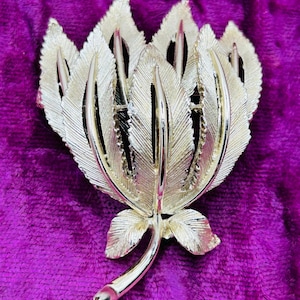 May include: A gold-tone metal brooch shaped like a cluster of textured, pointed leaves arranged in a fan-like formation. The piece features a central stem at the bottom with two small leaves attached, while the larger upper leaves have fine, etched lines.