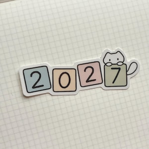 Puede incluir: Una pegatina troquelada que muestra el año 2027 en cuatro bloques cuadrados de colores pastel. Los bloques son azul claro, melocotón, rosa y verde salvia. Una pequeña ilustración de un gato blanco se sitúa sobre el bloque verde con el siete.
