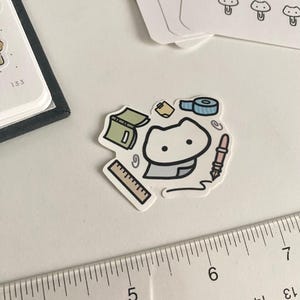 Puede incluir: Una pegatina con un personaje de dibujos animados rodeado de artículos de papelería. La pegatina incluye una regla, un libro, un rollo de cinta adhesiva, un bolígrafo y un bloc de notas. Se ven pegatinas con forma de seta en el fondo.