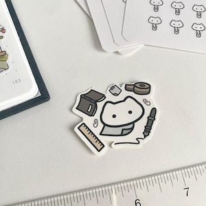 Puede incluir: Una pegatina con un gato de dibujos animados rodeado de artículos de papelería como una regla, un bolígrafo, cinta adhesiva y un cuaderno. También se ven pegatinas con forma de hongo y un cuaderno. La pegatina tiene un borde blanco.