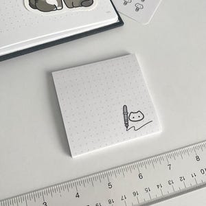 May include: A white square sticky note with a dotted grid pattern and a black pen and cat doodle. A clear ruler with black markings is in the foreground. Two gray cat stickers are on a notebook in the background.