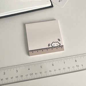 May include: A square notepad with a light brown border featuring a cartoon cat peeking over a ruler. The notepad is on a white surface with a clear ruler and stickers of cats and mushrooms. The notepad is approximately 3 inches wide.