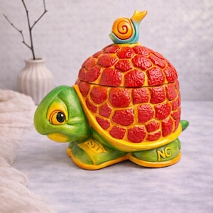 Könnte beinhalten: Ein keramikschildkrötenförmiger Behälter mit einem roten, strukturierten Panzer und einem grünen Kopf und Füßen. Auf dem Deckel sitzt eine kleine Schneckenfigur. Der Behälter hat gelbe Akzente und steht auf einer hellen Oberfläche.