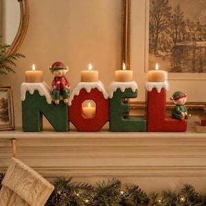 May include: A festive candle holder shaped like the word NOEL in red and green with white snow-like accents on top. The letters feature lit pillar candles, and two small elf figurines are perched on the letters N and L. The center O holds a candle.