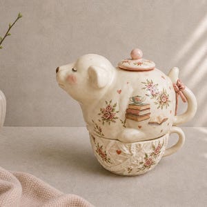 Op de afbeelding: Crèmekleurige keramische theepot en kop en schotel in de vorm van een hond. De theepot heeft een bloemmotief met roze rozen, harten en boeken. De kop en schotel heeft een bijpassend bloemmotief.