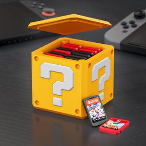 Può includere: Un contenitore di stoccaggio giallo a forma di blocco con punto interrogativo con un coperchio aperto, riempito di cartucce di gioco nere e rosse. Due cartucce di gioco Nintendo Switch sono davanti al contenitore. Una console Nintendo Switch è sullo sfondo.