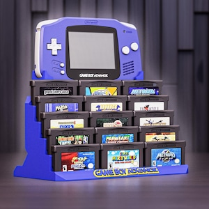 May include: A blue Game Boy Advance display stand with multiple game cartridges. The stand has a Game Boy Advance console design at the top and the words "GAME BOY ADVANCE" at the base. The cartridges are labelled with game titles.