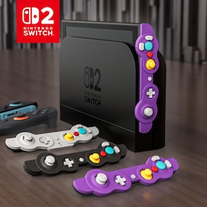 Puede incluir: Una consola Nintendo Switch negra con varios accesorios de mando de diferentes colores. Los mandos son negros, blancos y morados, con botones coloridos. El logotipo de Nintendo Switch es visible en la esquina superior izquierda.