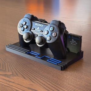 Könnte beinhalten: Schwarzer PlayStation 2-Controller und 8MB-Speicherkarte auf einem schwarzen Ständer. Der Controller hat das Sony PlayStation-Logo und verschiedene Tasten. Der Ständer hat blaue Akzente und einen Steckplatz für die Speicherkarte.