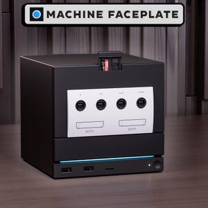 以下が含まれることがあります： 黒い立方体の電子機器。白いフェイスプレートには4つのコントローラーポートと「SLOT-A」「SLOT-B」とラベル付けされた2つのメモリーカードスロットがある。上部にはメモリーカードが挿入されており、底部には2つのUSBポートとライトストリップが備わっている。